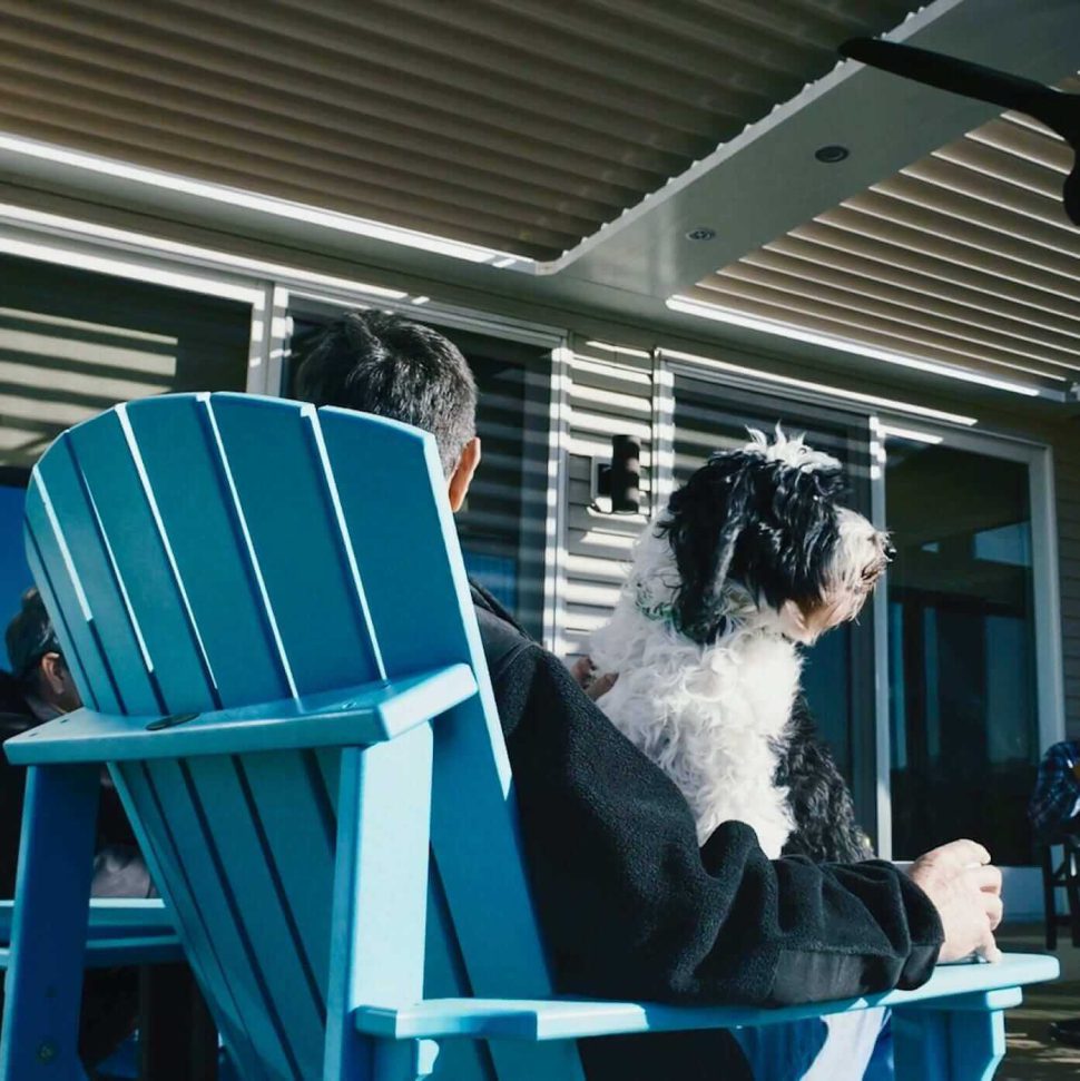Man in blue Adirondak chair holding black and white dog on lap under louvered roof outdoor structure