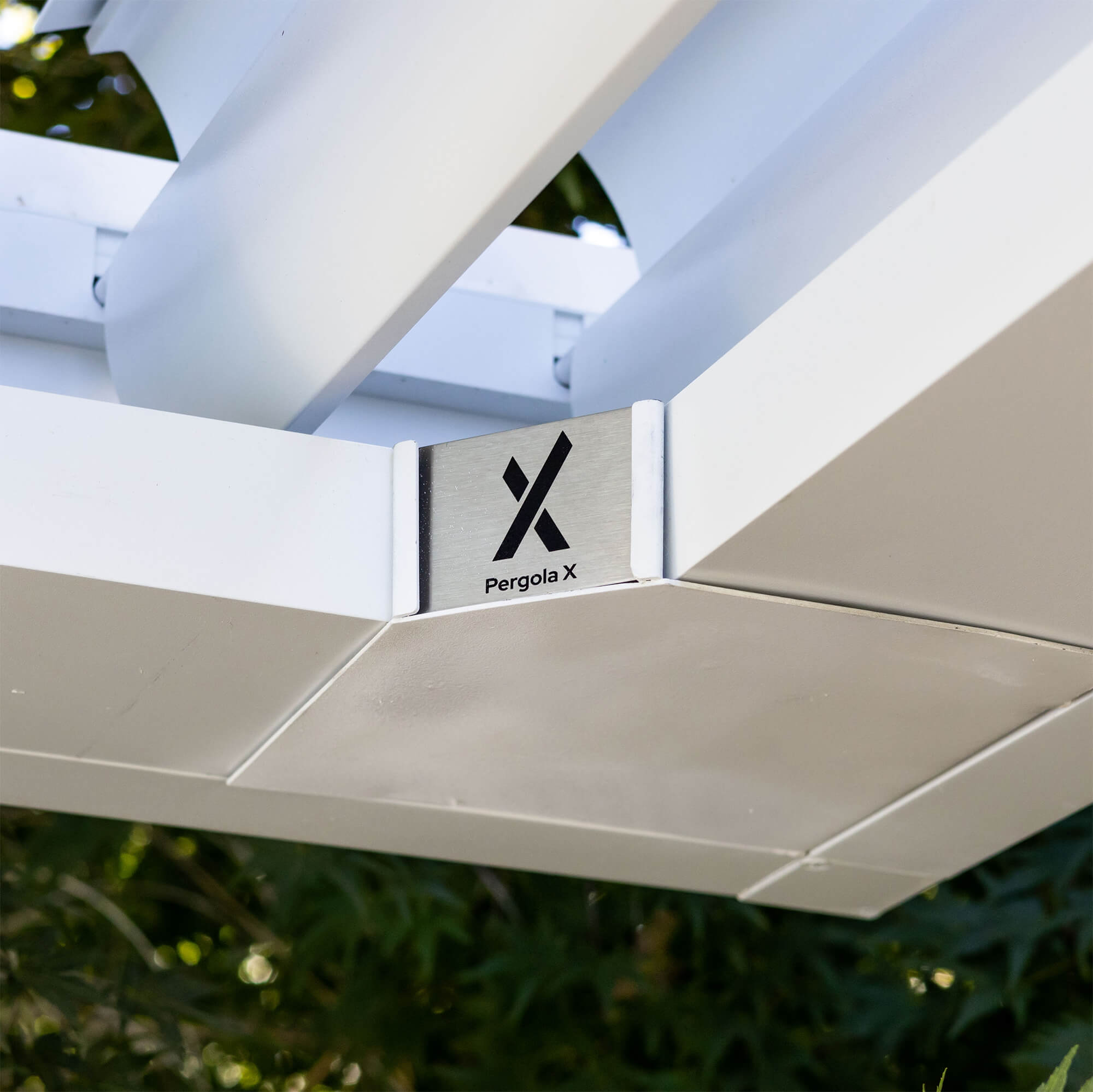 Close-up view of white aluminum Pergola X with modern louvered roof system and branded metal corner plate, set against a natural green background.