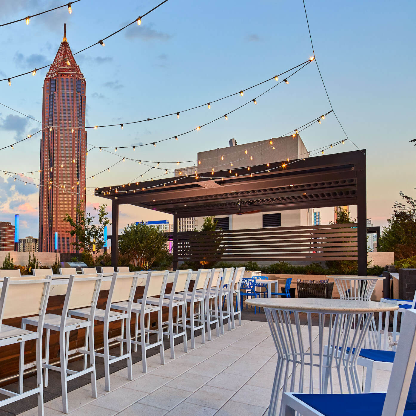 Modern rooftop patio with stylish louvered pergola, string lights, white bar seating, and city skyline view featuring the Bank of America Plaza in Atlanta at sunset.