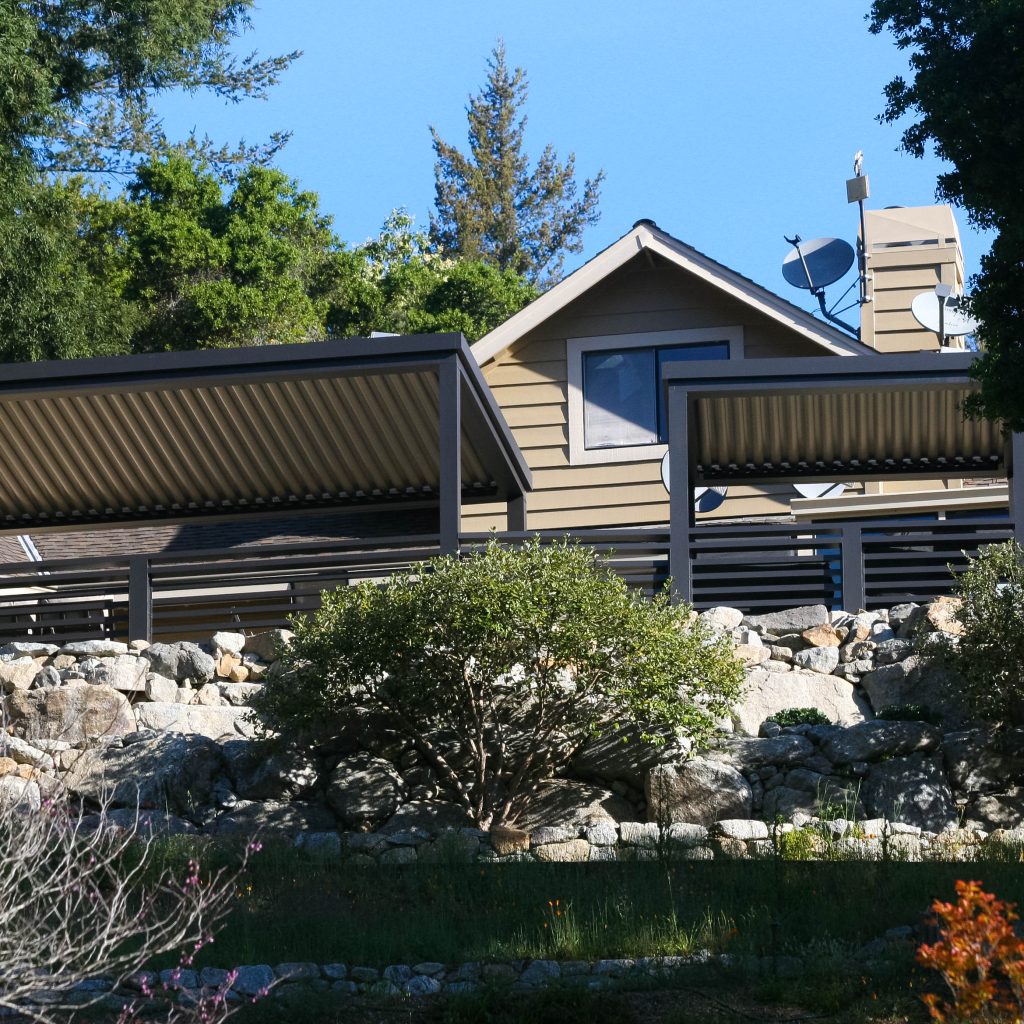 Modern hillside home with tan siding, multiple satellite dishes, and metal awnings, surrounded by stone landscaping and lush green trees under a clear blue sky.