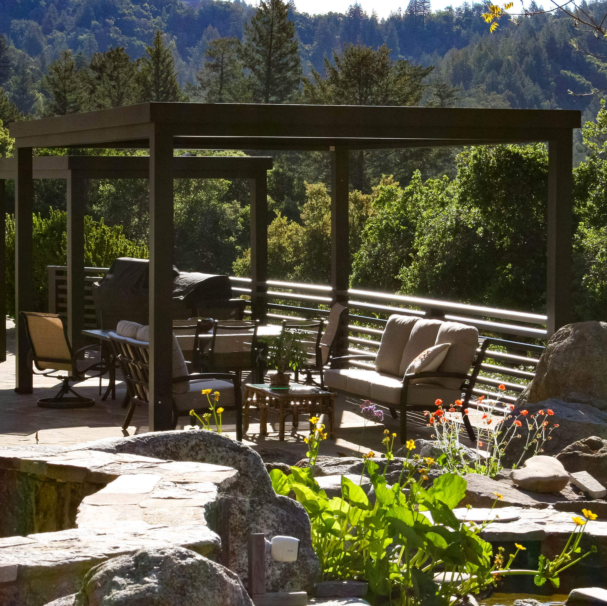 Modern outdoor patio with pergola, cushioned seating, and mountain forest views surrounded by lush greenery and garden landscaping