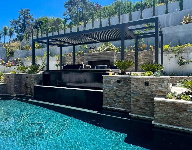 Luxury backyard with modern pergola lounge, stone waterfall feature, and infinity-edge pool surrounded by lush hillside landscaping