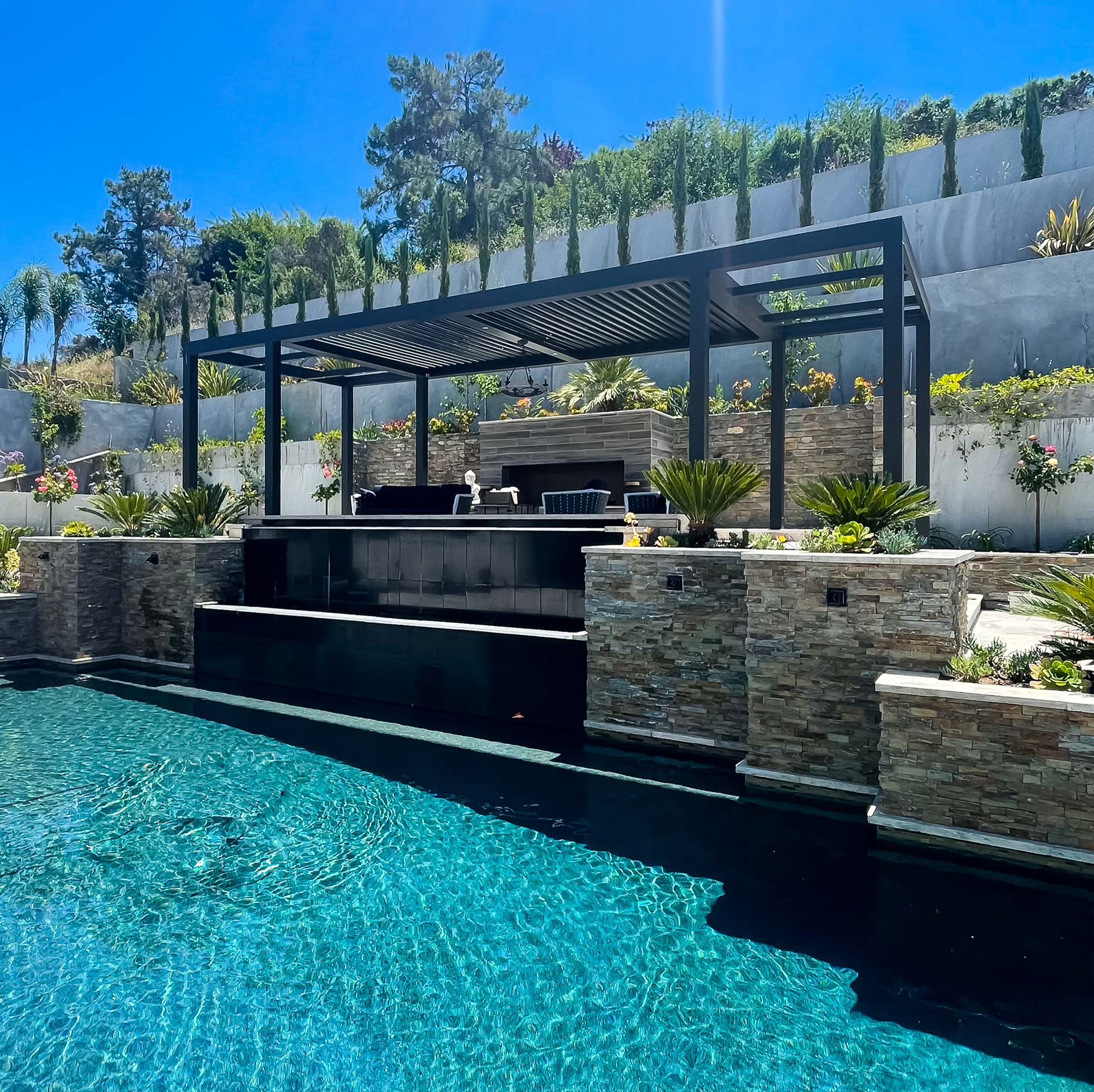 Luxury backyard with modern pergola lounge, stone retaining walls, and sparkling swimming pool surrounded by lush landscaping