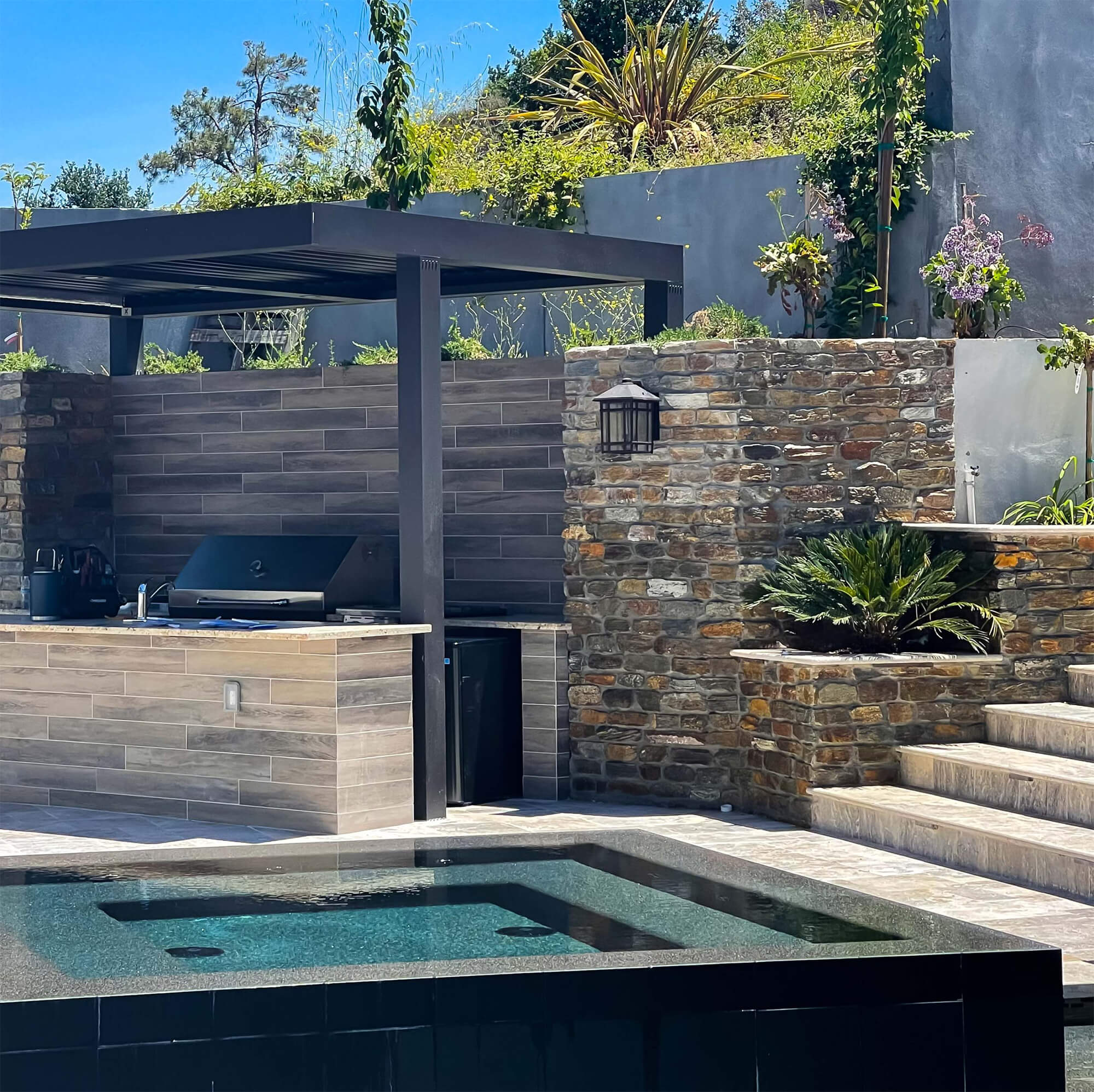 Modern outdoor kitchen with built-in grill and pergola next to a luxury spa pool and stone patio landscaping.